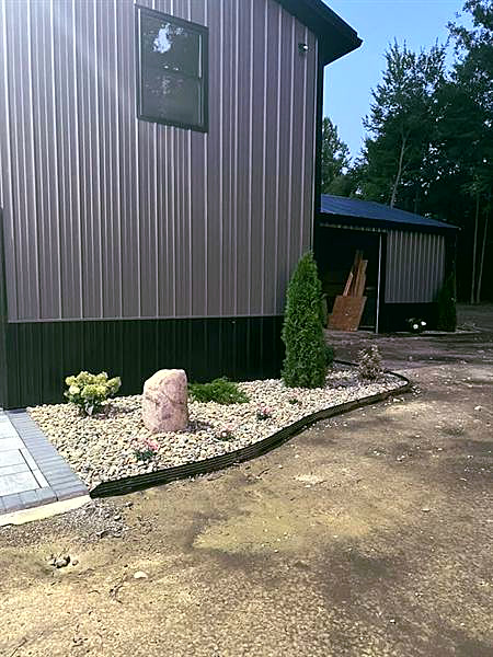 A rock bed with a boulder and arborvitae beside a pole barn, with bare dirt in front of it.