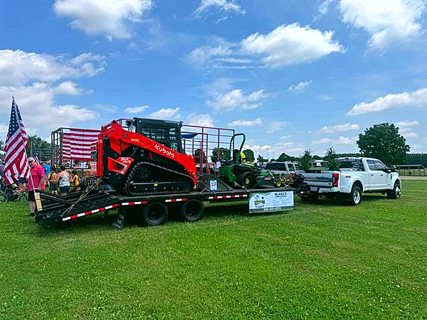 Blake's crew on a flatbed trailer with a skid steer and zero-turn mower, flags flying at a community parade.