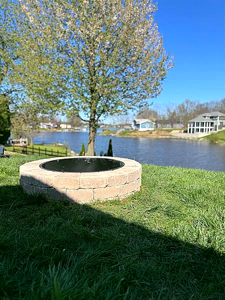 A stone fire pit on a lakeside lawn under a young tree on a clear day.
