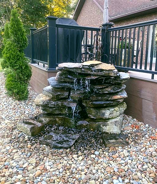 A stacked stone waterfall feature on river rock beside a deck with evergreens behind it.